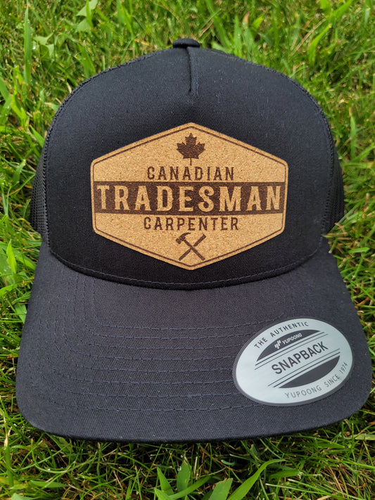 A black trucker hat with a brown patch on the front showing the text 'CANADIAN TRADESMAN CARPENTER' and a Canadian maple leaf symbol, placed on a grassy surface.