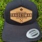 A black trucker hat with a brown patch on the front showing the text 'CANADIAN TRADESMAN CARPENTER' and a Canadian maple leaf symbol, placed on a grassy surface.