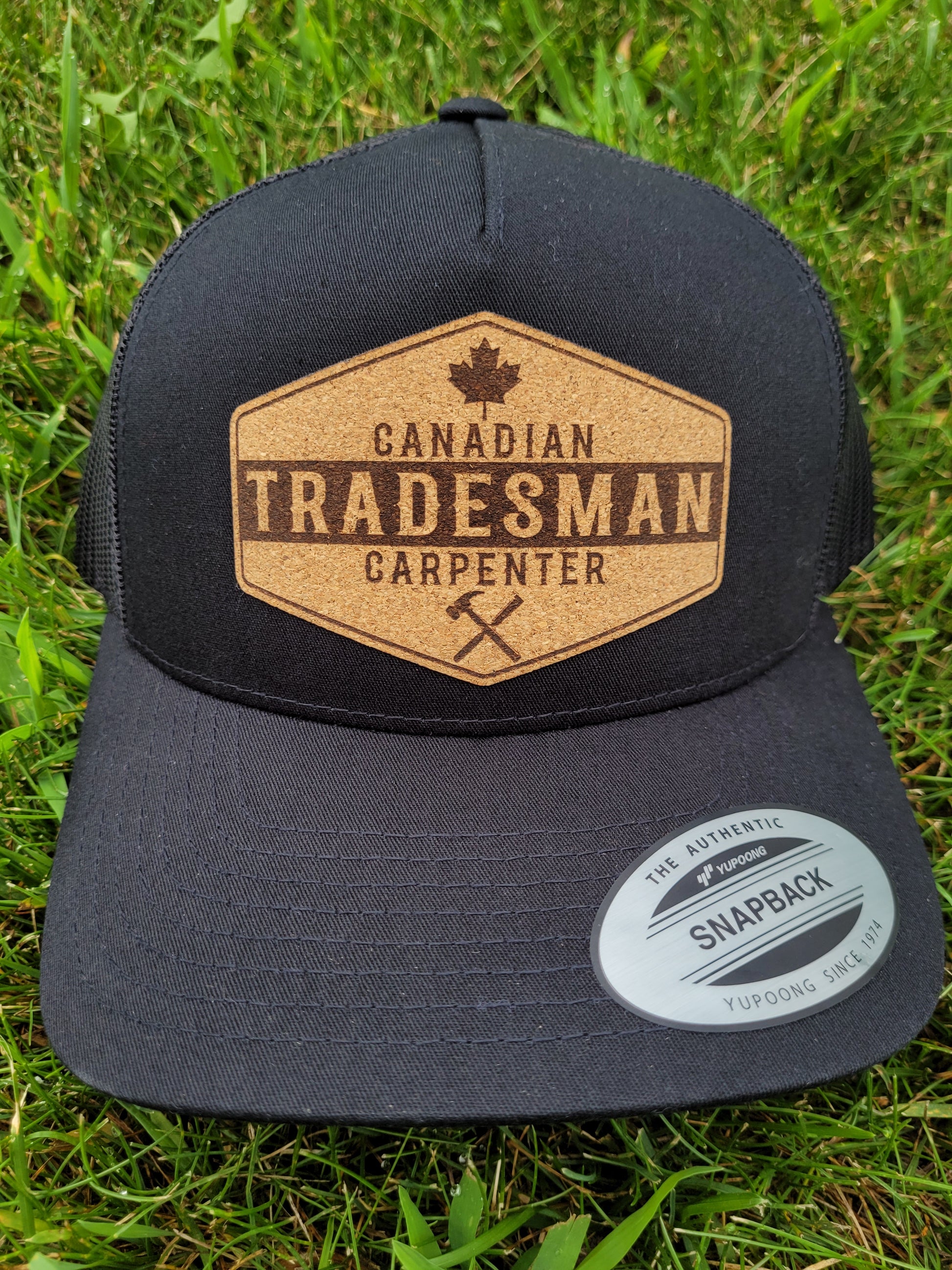 A black trucker hat with a brown patch on the front showing the text 'CANADIAN TRADESMAN CARPENTER' and a Canadian maple leaf symbol, placed on a grassy surface.