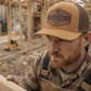 Man wearing a 'Canadian Tradesman' cap and overalls holding a piece of wood on a construction site.