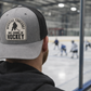 Person wearing a cap with 'Weekend Forecast 100% Chance of Hockey' text, looking at an ice hockey rink.